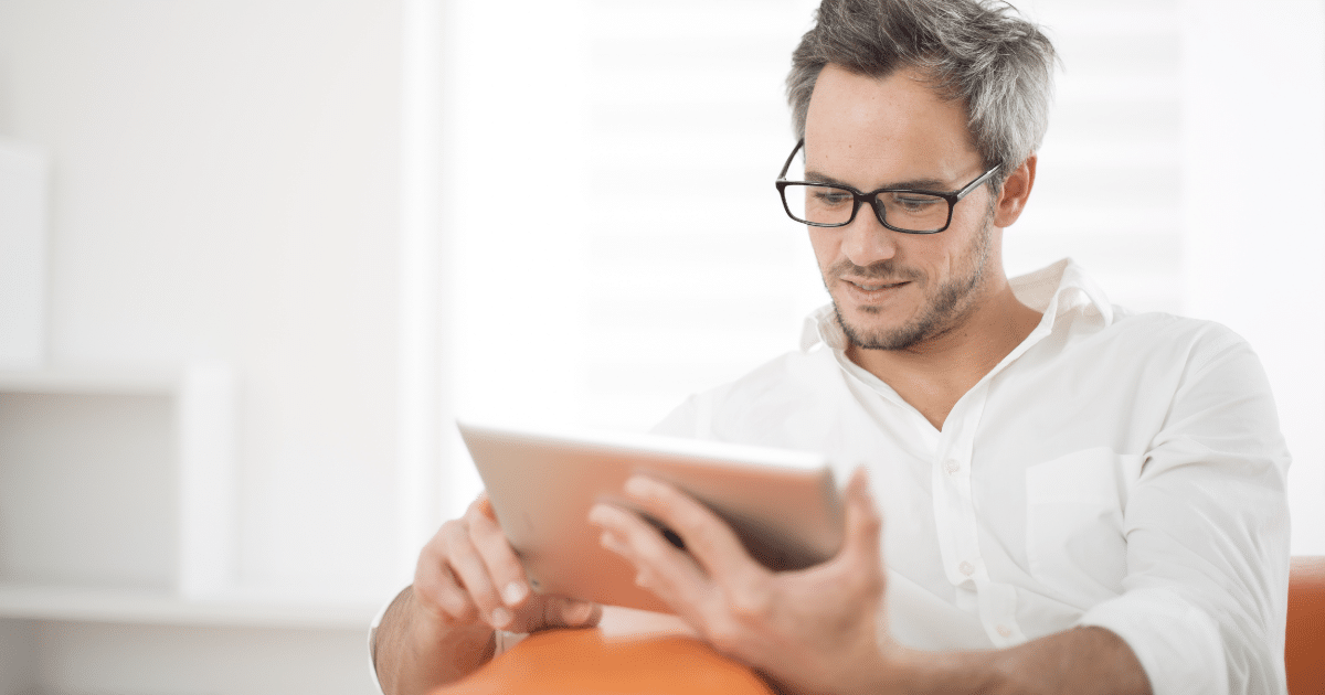 man sitting on couch reading on his tablet with glasses on